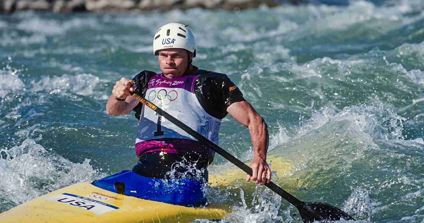 Male whitewater kayaker wearing a USA helmet and jersey paddles through rough water in a yellow kayak, gripping a black paddle.