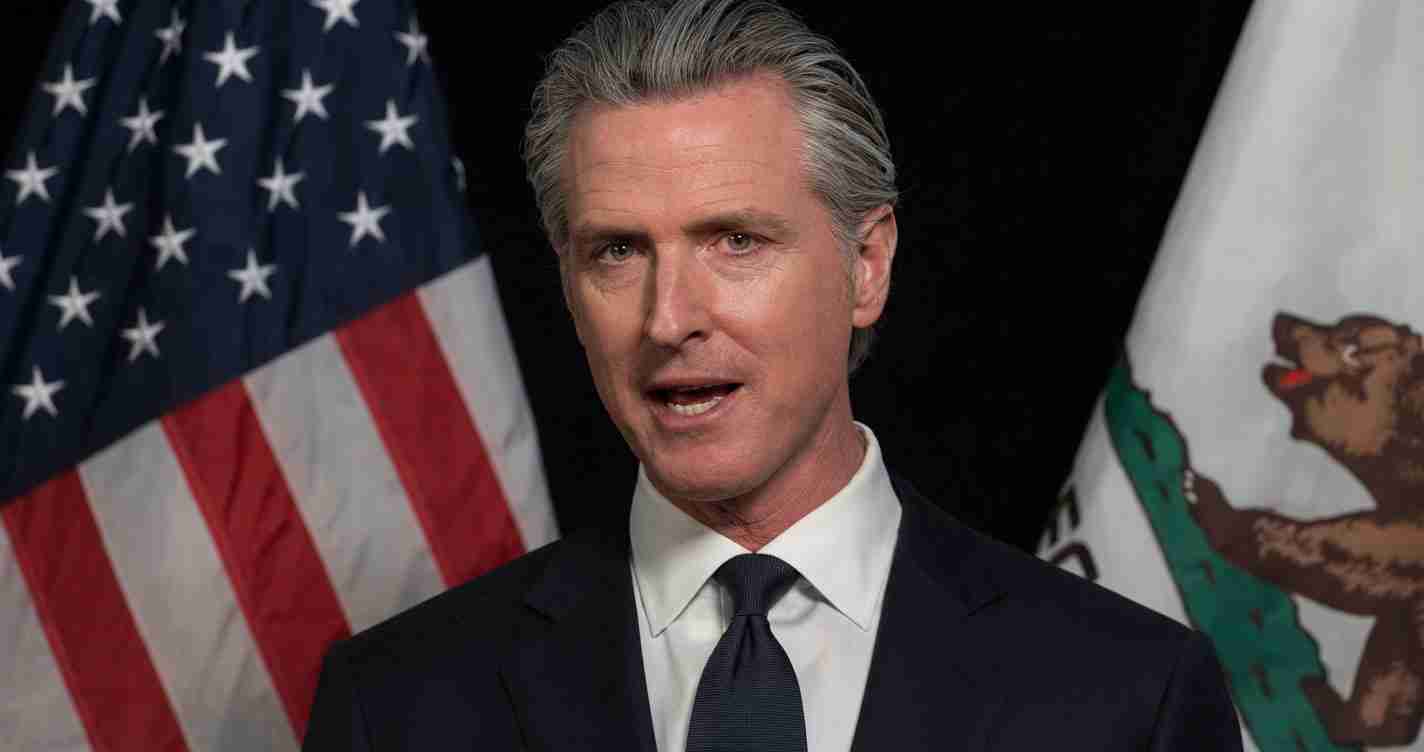 Politician in a dark suit speaking with the U.S. flag and a state flag behind him.