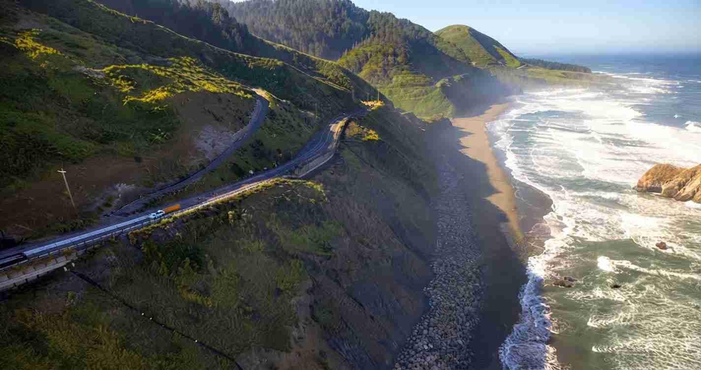 Coastal highway winding along green cliffs with the ocean to the right and a rocky shoreline. vehicles travel along the road.