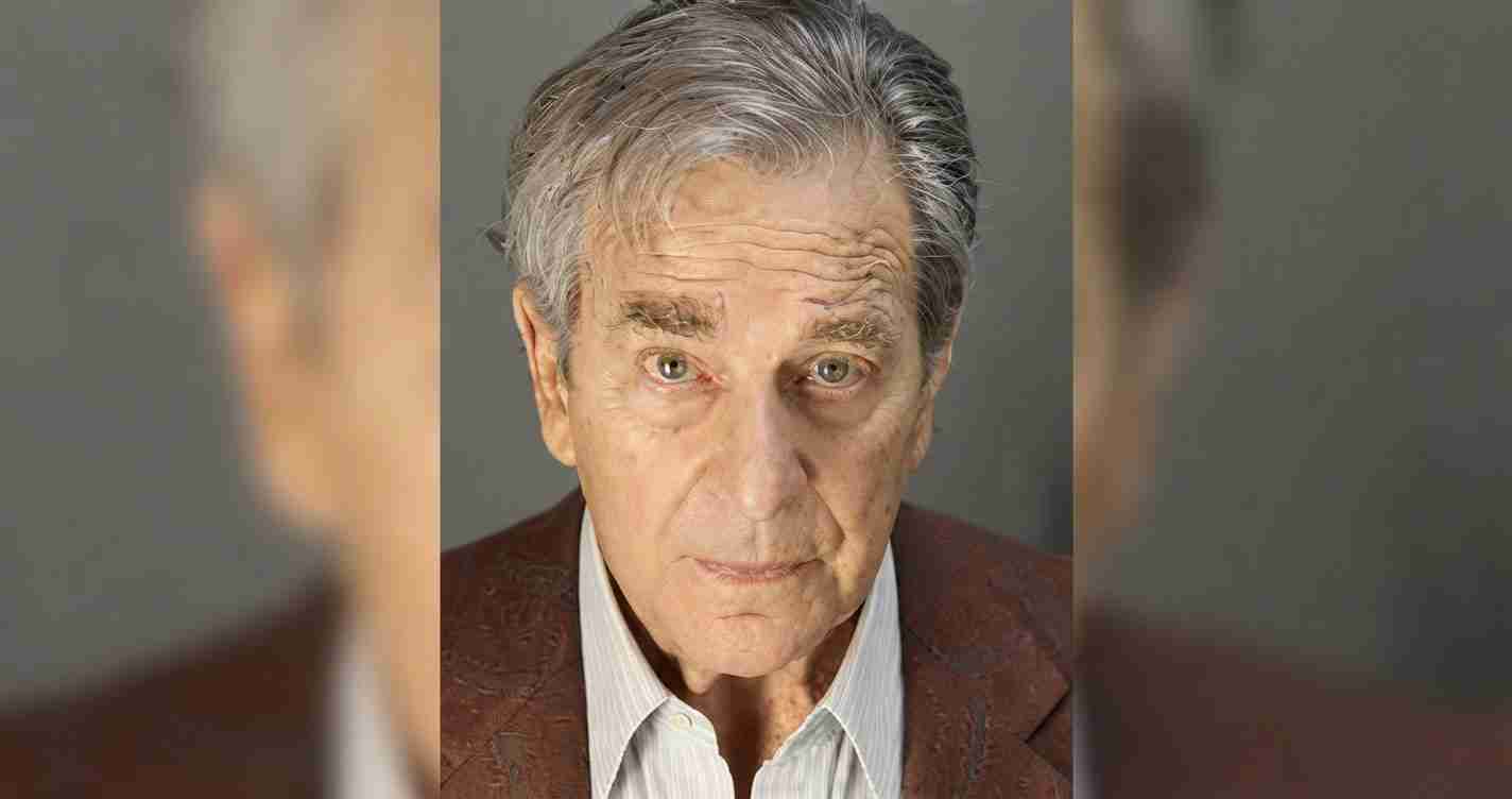 Close-up portrait of an older man with gray hair and a serious expression, wearing a brown blazer and light shirt.