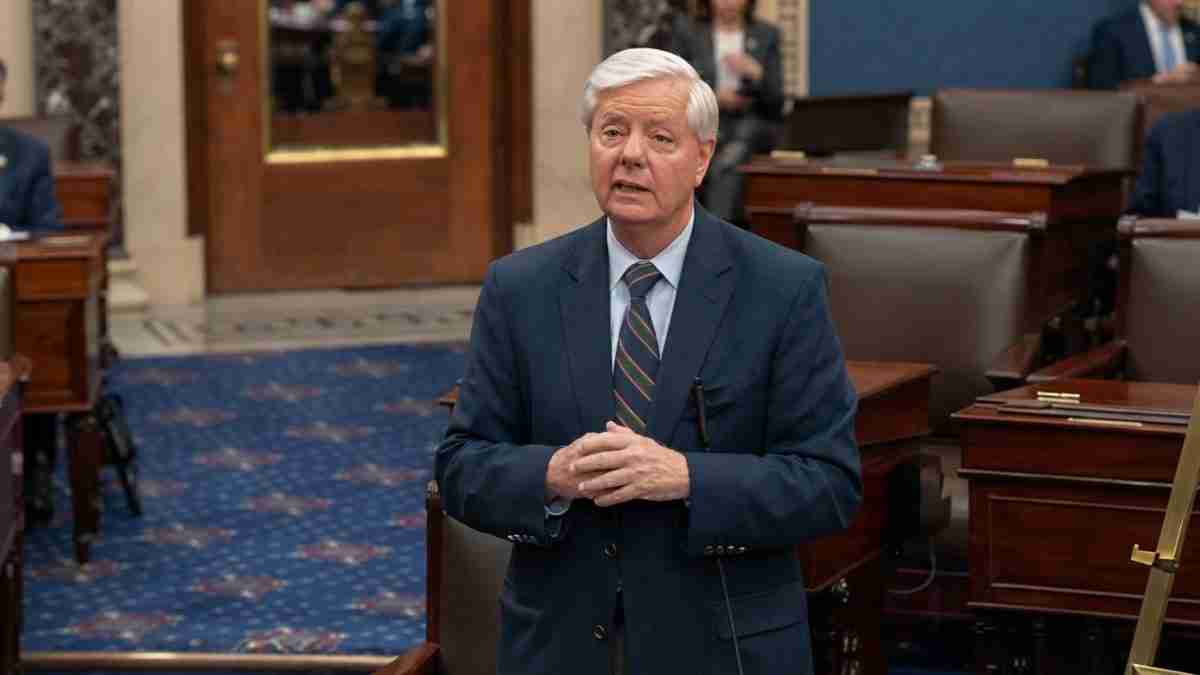 Politician in a navy suit speaks in a legislative chamber, hands clasped as he addresses colleagues.