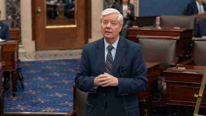 Politician in a navy suit speaks in a legislative chamber, hands clasped as he addresses colleagues.