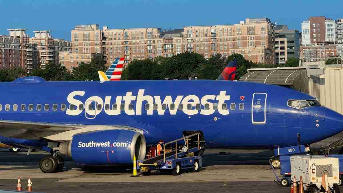 Blue Southwest airplane parked at a gate with boarding stairs and city buildings in the background.