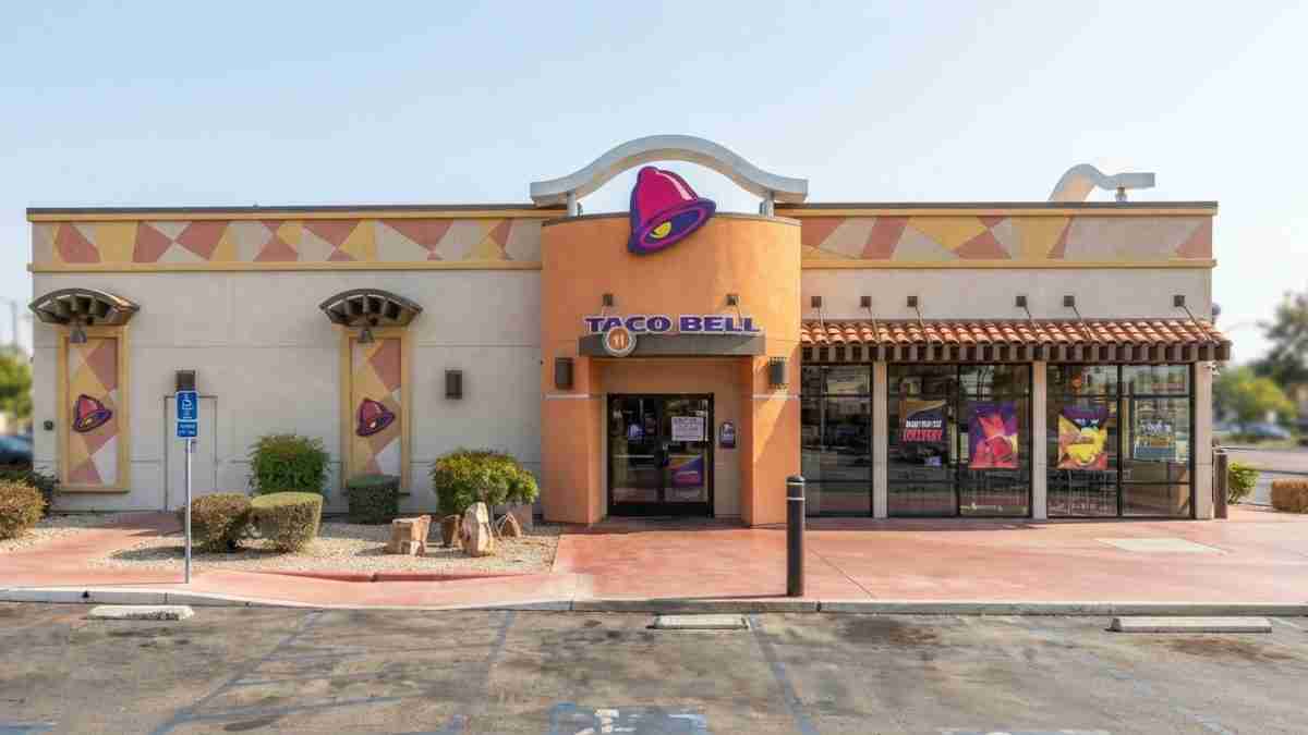 Exterior view of a Taco Bell restaurant with an orange tower and purple bell logo, beige walls, and large front windows displaying posters; parking lot in foreground.