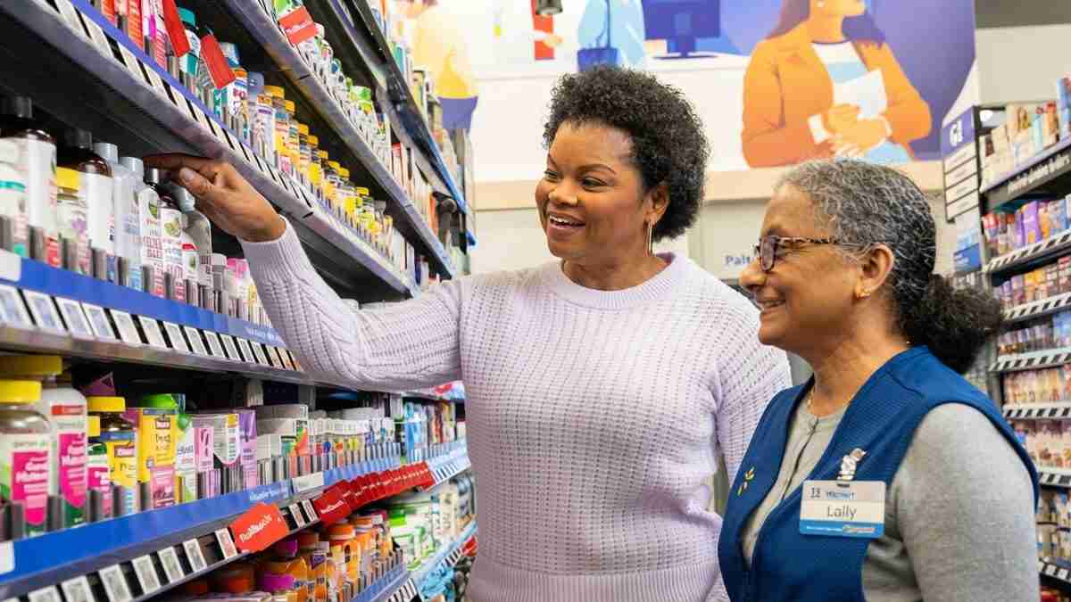 Two women in a drugstore aisle examine bottles on a shelf; one employee in a blue vest chats with a customer holding a product.