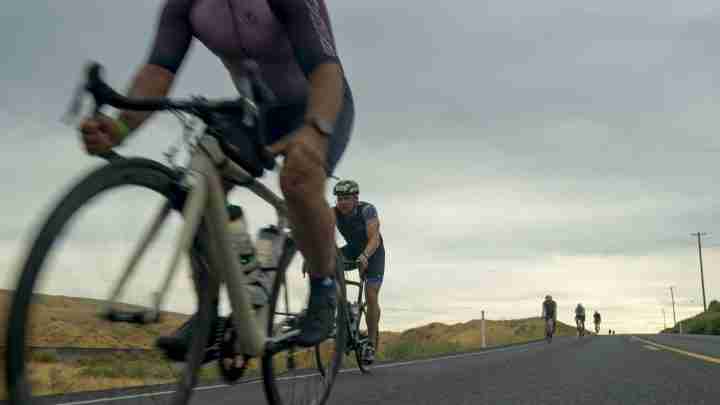 Cyclists ride on a rural road, foreground blur of a lead cyclist on a beige bike, scene of group cycling under cloudy sky.