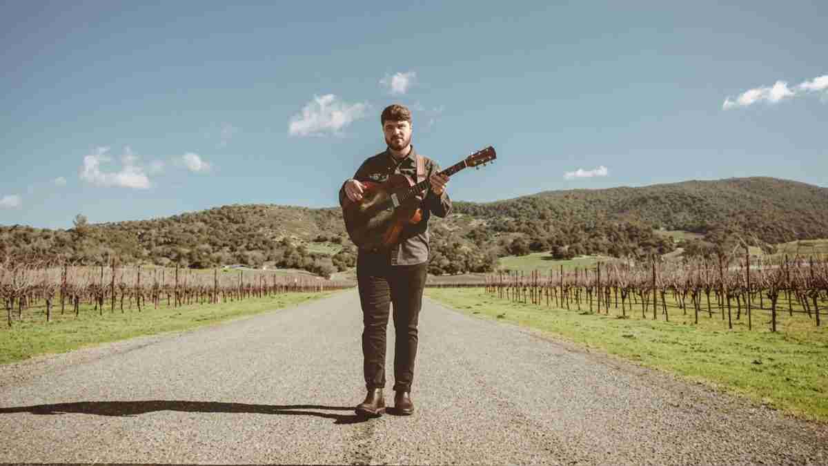 Man standing on a rural road with an acoustic guitar, brown boots, and hills in the background under a clear blue sky; rows of grapevines line the road on both sides.