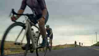 Cyclists riding on a straight rural highway under a cloudy sky; a rider in the foreground is blurred, others ahead on the road.