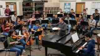 School orchestra rehearsal in a music room: conductor directs string, woodwind, and brass players while a pianist accompanies at a grand piano.