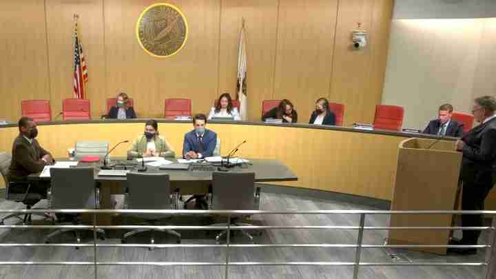 Panel discussion in a government chamber: five people sit at a curved table, two speakers at the front, California seal and flags behind them.